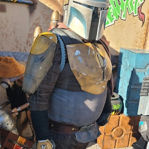 May include: A person dressed as a Mandalorian, wearing a blue and gray helmet with a yellow and brown chest plate and a brown belt. The person is standing in front of a wall with graffiti and a brown box.