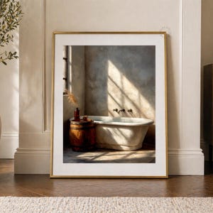 May include: A framed print depicts a vintage bathroom scene. A white clawfoot bathtub is positioned in front of a window, with sunlight streaming in. A small wooden barrel holds bottles and other items. The frame is gold.