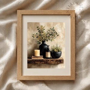 May include: A framed wall art print featuring a still life composition on a wooden shelf. The scene includes a black vase with green foliage, a beige pillar candle on a book, a black pump bottle, and a small black bowl with green plants.