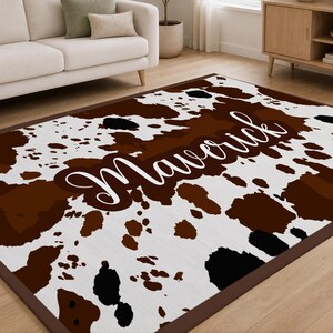 May include: A rectangular area rug featuring a cowhide pattern with irregular patches of dark brown, black, and white. The center of the rug displays the name Maverick in a white cursive script font. The rug has a solid dark brown border.