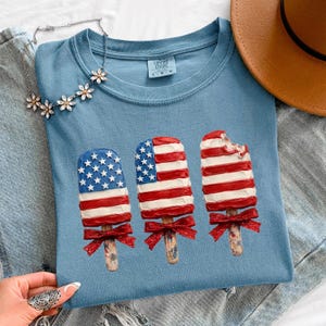 Può includere: Una t-shirt blu con la grafica di tre ghiaccioli decorati con motivi della bandiera americana in rosso, bianco e blu. Ogni ghiacciolo ha un fiocco rosso glitterato legato allo stecco di legno. La maglietta ha un'etichetta del marchio Comfort Colors.