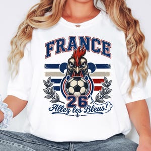 May include: White t-shirt featuring a graphic of a rooster with a soccer ball, the word "FRANCE" in blue, and the text "Allez les Bleus!" in blue. The design incorporates the colors red, white, and blue, with a distressed vintage style.