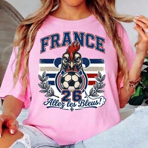 May include: Pink t-shirt with a graphic featuring the word "FRANCE" in blue and red, a rooster head, a soccer ball, the number "26", and the phrase "Allez les Bleus!" in blue. The design includes stripes and laurel branches.