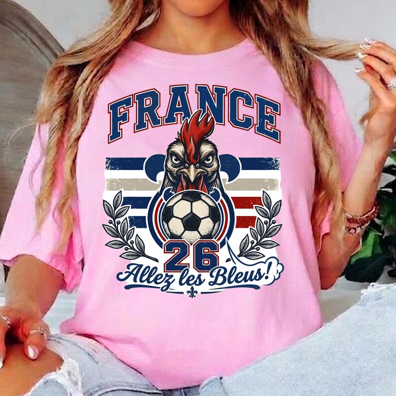May include: Pink t-shirt with a graphic featuring the word "FRANCE" in blue and red, a rooster head, a soccer ball, the number "26", and the phrase "Allez les Bleus!" in blue. The design includes stripes and laurel branches.