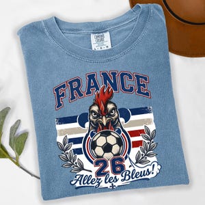 May include: Light blue t-shirt with a graphic featuring a rooster holding a soccer ball, the word "FRANCE" in blue, and the text "Allez les Bleus!" in white. The shirt has a crew neck and short sleeves.