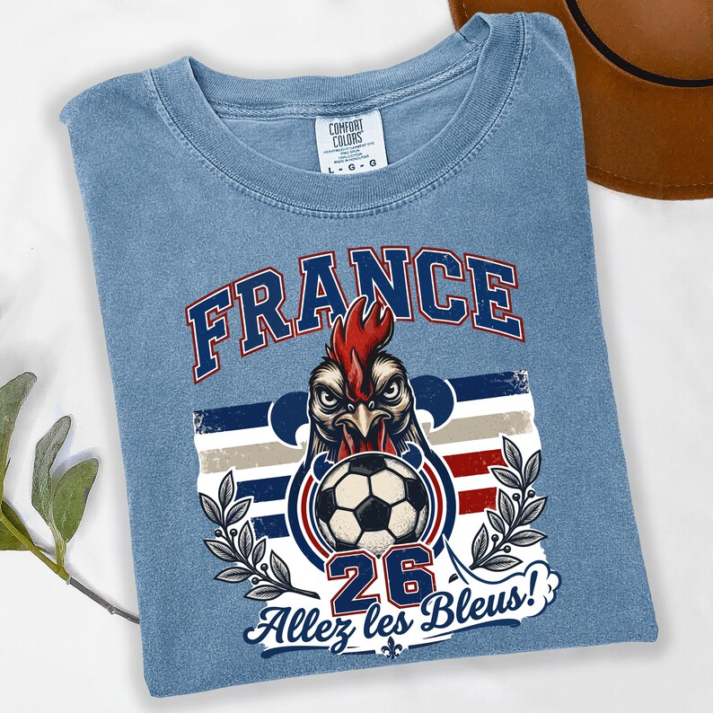 May include: Light blue t-shirt with a graphic featuring a rooster holding a soccer ball, the word "FRANCE" in blue, and the text "Allez les Bleus!" in white. The shirt has a crew neck and short sleeves.