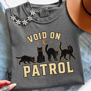 Może przedstawiać: Ciemnoszary t-shirt z grafiką przedstawiającą cztery czarne koty w różnych pozach oraz napisem "VOID ON PATROL" kremową czcionką. Nad kotami znajduje się mały pomarańczowy półksiężyc i dwie gwiazdki. Koszulka ma okrągły dekolt.