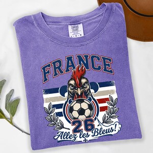 May include: A purple t-shirt with a graphic featuring a rooster holding a soccer ball. The word "FRANCE" is arched across the top in blue and white. Below the rooster is the number "26" and the phrase "Allez les Bleus!"