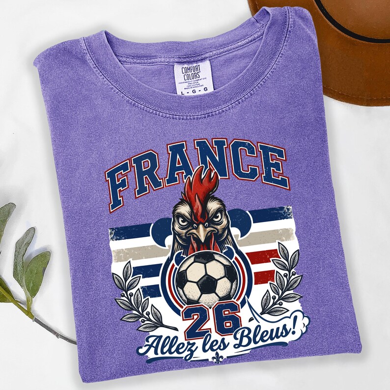May include: A purple t-shirt with a graphic featuring a rooster holding a soccer ball. The word "FRANCE" is arched across the top in blue and white. Below the rooster is the number "26" and the phrase "Allez les Bleus!"