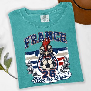 May include: Teal Comfort Colors t-shirt featuring a graphic of a rooster with a soccer ball, the word "FRANCE", the number "26", and the phrase "Allez les Bleus!". The design incorporates the colors of the French flag.
