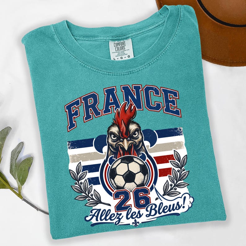 May include: Teal Comfort Colors t-shirt featuring a graphic of a rooster with a soccer ball, the word "FRANCE", the number "26", and the phrase "Allez les Bleus!". The design incorporates the colors of the French flag.