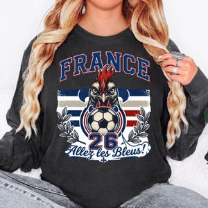 May include: Dark gray sweatshirt featuring a graphic of a rooster with a soccer ball, the word "FRANCE" in blue, and the text "26 Allez les Bleus!" in white. The design incorporates the colors of the French flag.