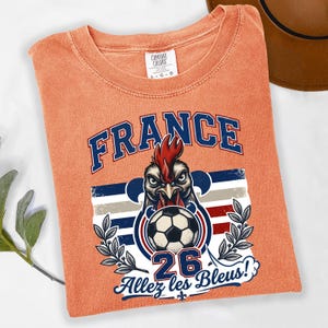 May include: Orange Comfort Colors t-shirt featuring a graphic of a rooster holding a soccer ball, with the word "FRANCE" above and "26 Allez les Bleus!" below. The shirt has a crew neck and short sleeves.