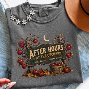 May include: A charcoal gray t-shirt featuring a graphic of a wooden sign that reads "AFTER HOURS AT THE ORCHARD" with the text "NIGHT PICKING" and "AUTUMN SEASON" below. The design includes red apples, a crescent moon, and a wooden basket.