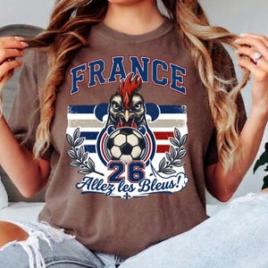May include: Brown t-shirt with a graphic featuring the word "FRANCE" in blue, a rooster with a soccer ball, and the text "Allez les Bleus!" in blue. The number "26" is also displayed. The shirt has a crew neck.