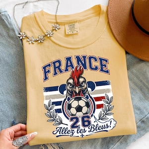 May include: A light yellow t-shirt featuring a graphic of a rooster holding a soccer ball, with the word "FRANCE" in blue. The shirt also includes the text "26" and "Allez les Bleus!"