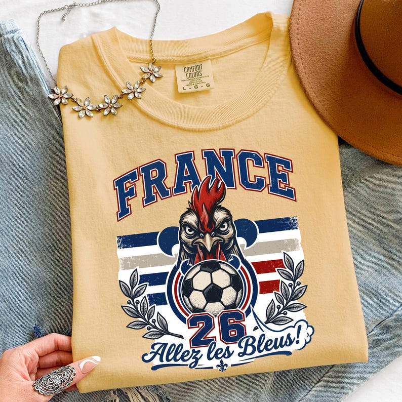 May include: A light yellow t-shirt featuring a graphic of a rooster holding a soccer ball, with the word "FRANCE" in blue. The shirt also includes the text "26" and "Allez les Bleus!"