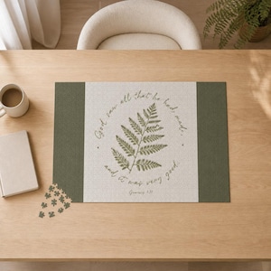 Puede incluir: Un rompecabezas rectangular que presenta un panel central de color crema con una ilustración de helecho verde y el texto 'God saw all that he had made, and it was very good. Genesis 1:31' en cursiva, flanqueado por bordes laterales verde oliva.