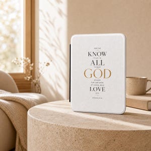 May include: A white rectangular e-reader case featuring the black and gold text: AND WE KNOW THAT IN ALL THINGS GOD WORKS FOR THE GOOD OF THOSE WHO LOVE HIM ROMANS 8:28. The case has a textured surface and a black spine on the left side.
