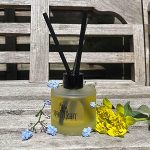 May include: A frosted glass aromatherapy diffuser with black reeds and a black cap. The bottle contains a yellow liquid and a label reading "Storylight Candle Co." Blue forget-me-nots and yellow flowers are arranged around the diffuser on a wooden surface.