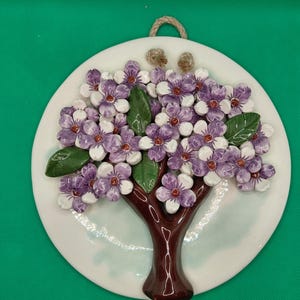 May include: A circular ceramic wall plaque featuring a three-dimensional tree with a dark brown trunk and numerous white and purple flowers. The tree has several green leaves and is mounted on a white background with a light blue wash.