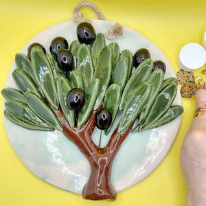 Puede incluir: Colgante de pared de cerámica con un olivo. El árbol presenta hojas verdes y aceitunas negras sobre un fondo circular blanco. El tronco es marrón y tiene una cuerda para colgarlo. Es un artículo decorativo.