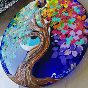 May include: A circular blue glass wall hanging featuring a hand-painted brown tree with textured bark and colorful leaves in shades of yellow, orange, pink, purple, and green. A large, central evil eye symbol with white, light blue, and black rings.