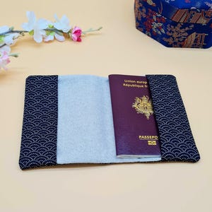 May include: Passport holder with a navy blue and white wave pattern on the exterior. The interior is light gray, and a burgundy passport is visible inside. A blue floral box is in the background.