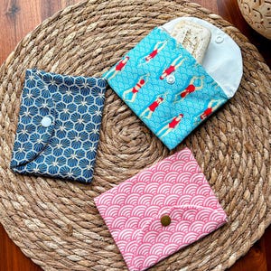 May include: Three fabric pouches with different patterns. The pouches are made of cotton and have a snap closure. The patterns are geometric, a blue and white pattern with swimmers, and a pink and white pattern with waves.