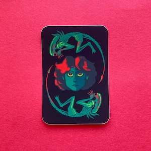 May include: A rectangular sticker with a dark background features a stylized illustration. A central figure with green skin and red hair is surrounded by two green alien figures. The sticker has rounded corners and is set against a pink background.