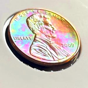 May include: A close-up of a 2009 US penny with a rainbow iridescent finish. The coin features the profile of Abraham Lincoln and the words "In God We Trust" and "Liberty".