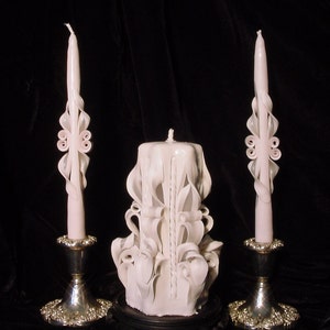 May include: Three white carved candles on silver candle holders. The center candle is larger and has a more elaborate design. The candles are on a black wooden base.
