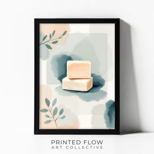 May include: A framed wall art print featuring two stacked rectangular beige soap bars centered against a background of soft watercolor washes in shades of blue, green, and peach, accented with delicate green leaf illustrations and the text PRINTED FLOW ART COLLECTIVE.