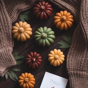 May include: A collection of seven small pumpkin-shaped candles in various colors, including orange, green, brown, and yellow, arranged on a wooden surface with a brown knitted sweater and green leaves. A card with the text "When you are some reason, we'll be happy!" is also on the surface.