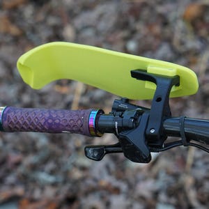 Puede incluir: Un protector de manillar amarillo con un soporte negro fijado a un manillar de bicicleta. El manillar tiene una empu&ntilde;adura morada y negra con un anillo de colores arco&iacute;ris.