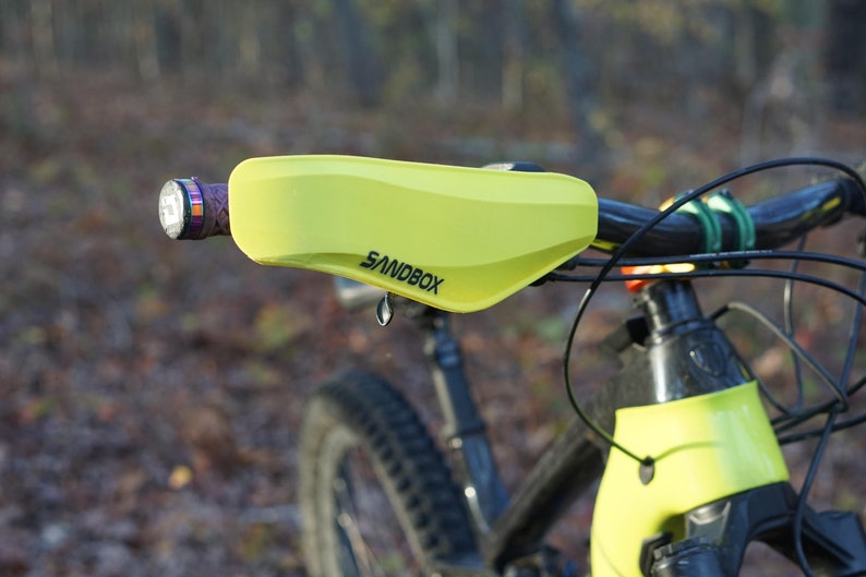 Puede incluir: Un protector de manillar amarillo con la palabra "SANDBOX" impresa en &eacute;l. El protector est&aacute; unido a un manillar de bicicleta de monta&ntilde;a negro.