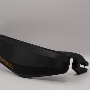 Puede incluir: Manillar de pl&aacute;stico negro con un logotipo "SANDBOX" dorado. El manillar tiene una forma curva y una abrazadera en el lado derecho.
