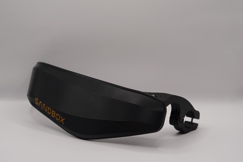 Puede incluir: Manillar de pl&aacute;stico negro con un logotipo "SANDBOX" dorado. El manillar tiene una forma curva y una abrazadera en el lado derecho.