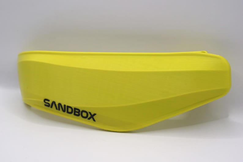 Puede incluir: Un casco amarillo brillante con la palabra "SANDBOX" impresa en negro en el lateral.