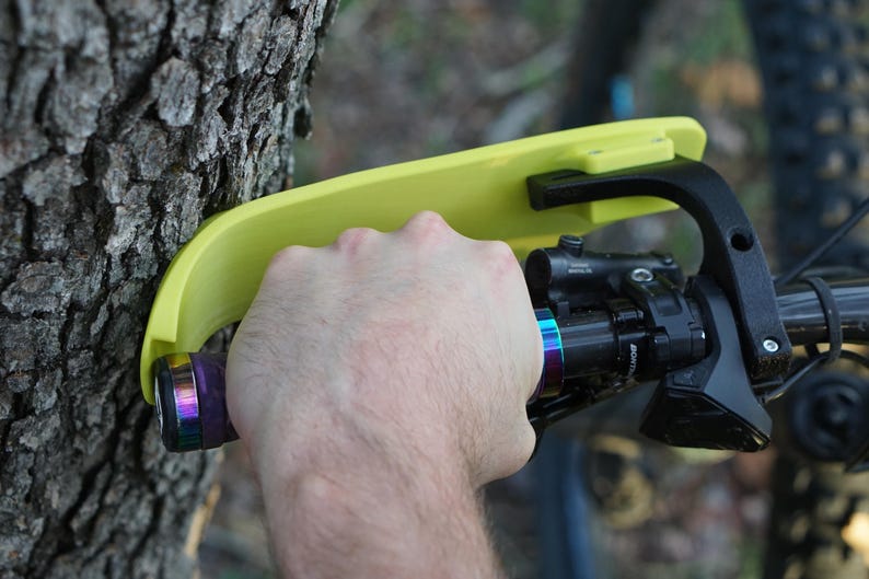 Puede incluir: Un soporte de manillar de bicicleta amarillo fijado a un tronco de &aacute;rbol. El soporte tiene un brazo negro y una palanca negra. El manillar de la bicicleta es negro con una empu&ntilde;adura de arco&iacute;ris.