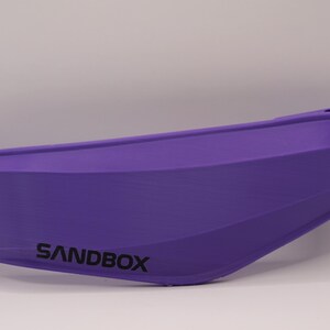 Puede incluir: Visera de casco de color morado con la palabra "SANDBOX" impresa.
