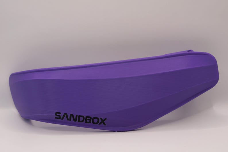 Puede incluir: Visera de casco de color morado con la palabra "SANDBOX" impresa.