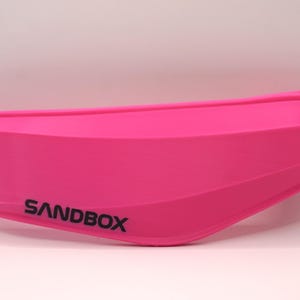 Puede incluir: Una visera de casco de pl&aacute;stico rosa con la palabra "SANDBOX" impresa en negro.