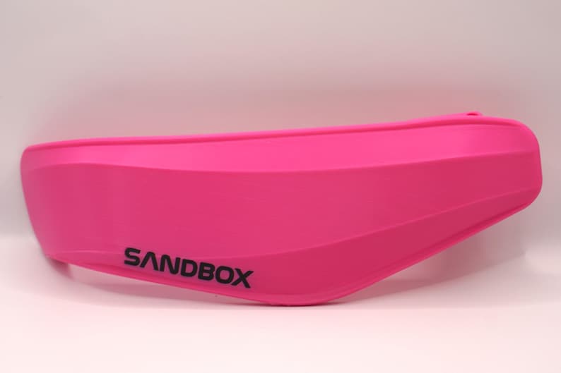 Puede incluir: Una visera de casco de pl&aacute;stico rosa con la palabra "SANDBOX" impresa en negro.