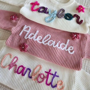Op de afbeelding: Drie gebreide truien met geborduurde namen in verschillende kleuren. De bovenste trui is wit met de naam "Taylor" geborduurd in roze, paars, blauw en groen garen. De middelste trui is roze met de naam "Adelaide" geborduurd in wit garen. De onderste trui is wit met de naam "Charlotte" geborduurd in roze, oranje en paars garen.