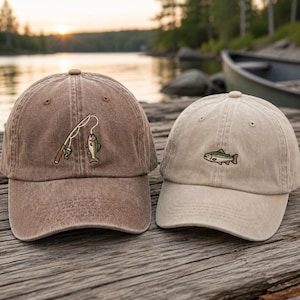 May include: Two baseball caps made of washed cotton fabric. The left cap is dark brown and features an embroidered fishing rod with a hooked fish. The right cap is light beige and features a single embroidered fish design on the front panel.
