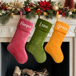 May include: Three velvet Christmas stockings in pink, olive green, and mustard yellow. Each stocking features a folded cuff with a name embroidered in white script: Harper on the pink, Lena on the green, and Grace on the yellow. Each has a loop.