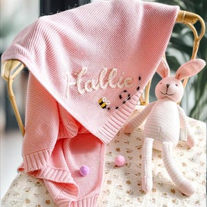 Peut inclure: Une couverture pour bébé en tricot rose avec le nom "Hallie" brodé dessus. Il y a une petite abeille brodée et un lapin en peluche sur la couverture.