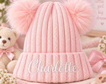 May include: A light pink ribbed knit beanie featuring two large, fluffy pom-poms on the top. The front of the folded cuff is embroidered with the name Charlotte in white cursive script. The hat has a vertical ribbed texture throughout the design.