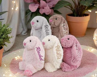 May include: Five plush stuffed bunnies in shades of gray, brown, pink, white, and mauve. Each bunny features long floppy ears with embroidered names including Rachel, Eulalia, and Candice. The soft toys have small pink noses and dark eyes.